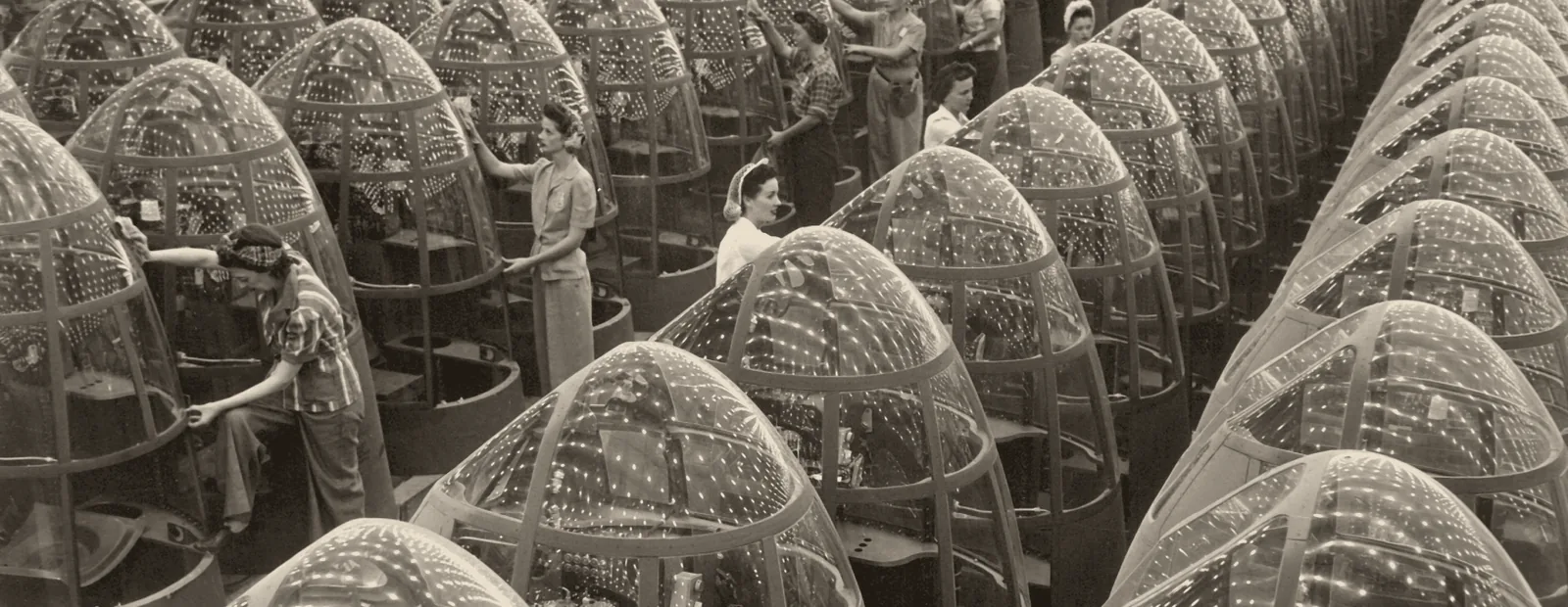 Rows of aircraft fuselage assemblies receding into the distance on a 1940s factory floor, workers between the lines