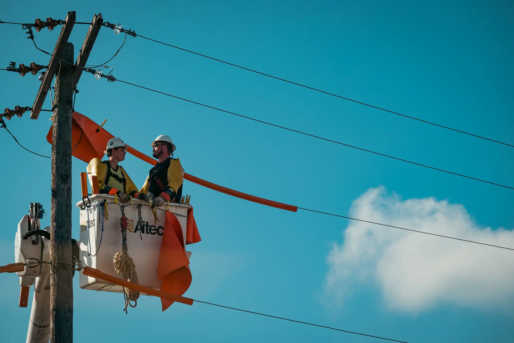 Two utility workers in hard hats and harnesses in an aerial lift beside power lines covered with rubber protection