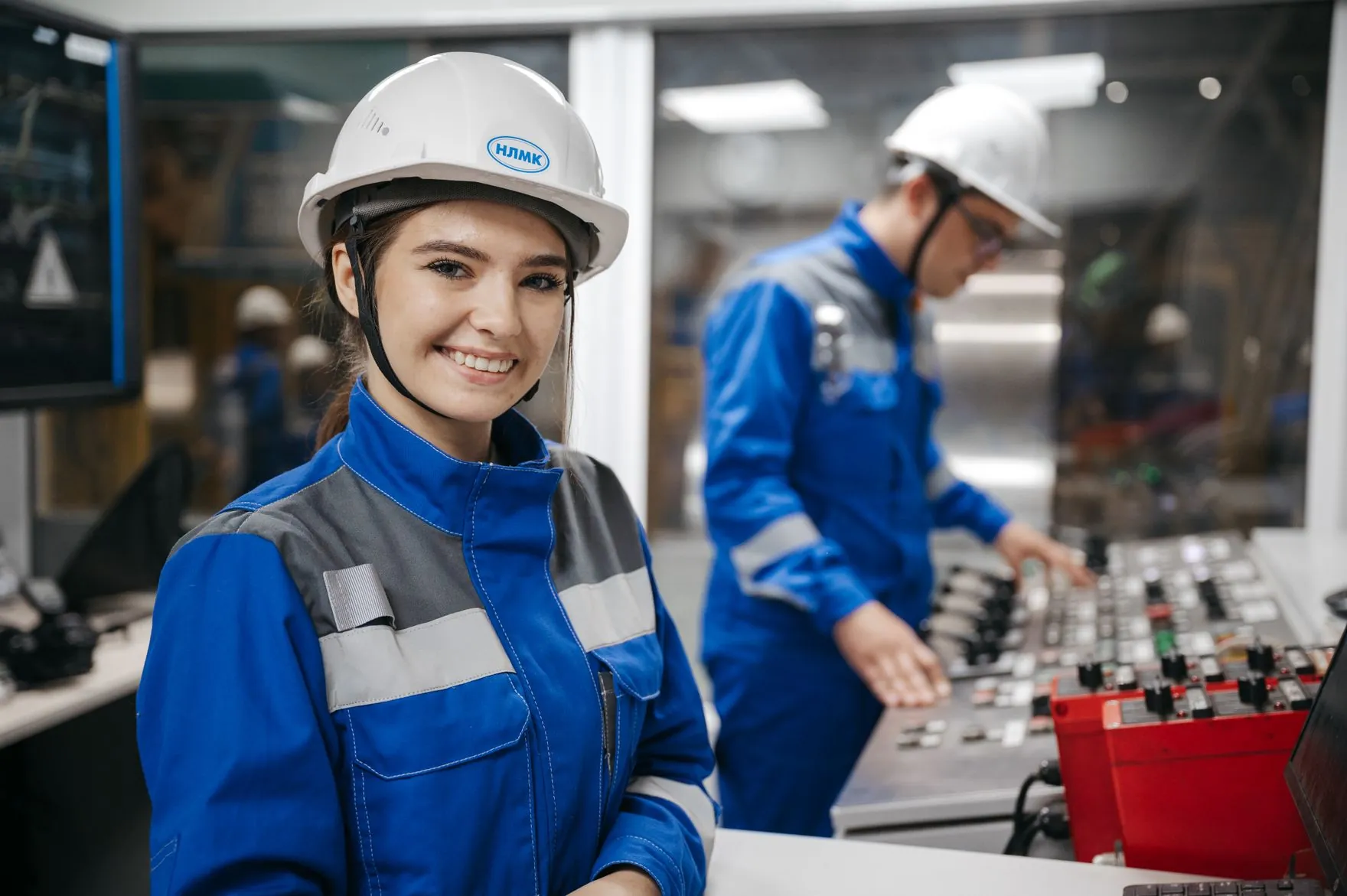 A smiling plant worker in a white hard hat and a blue coverall in a control room, a colleague at the panel behind her