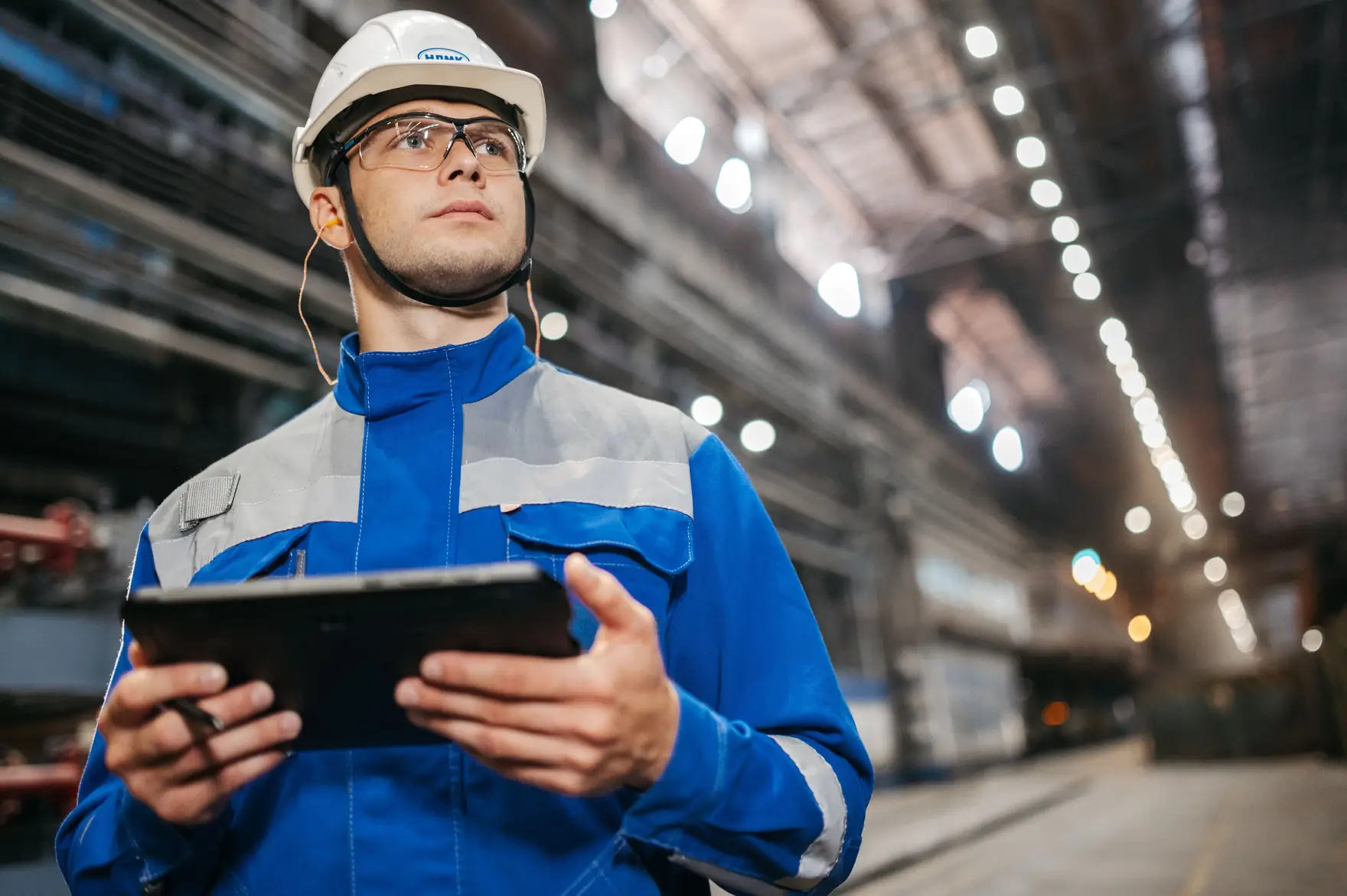 A plant worker in a hard hat, safety glasses and a blue coverall holding a tablet in a factory hall