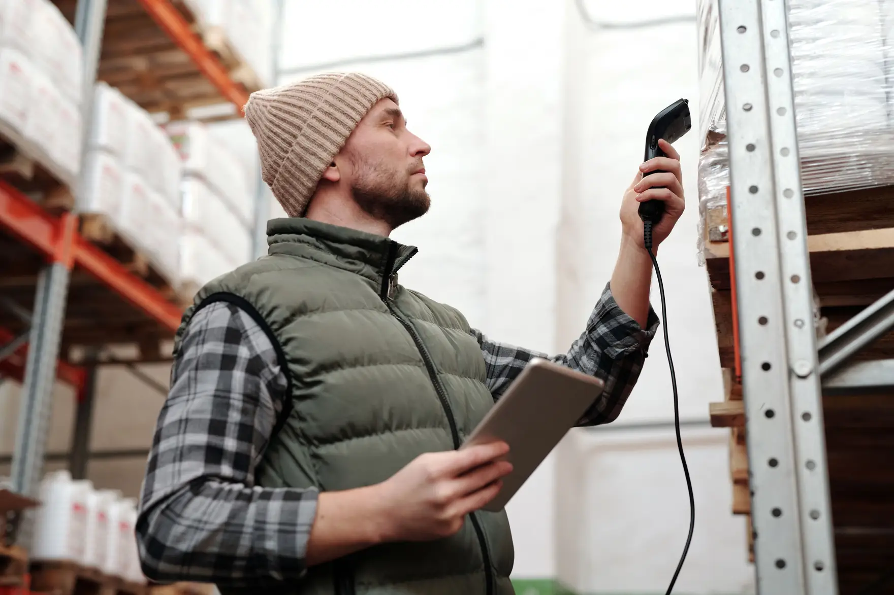 A warehouse worker in a vest scanning a pallet label on a rack while holding a tablet